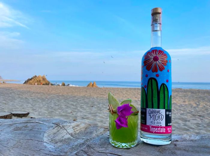 A bottle of mezcal next to a vibrant green cocktail set against a beach backdrop.