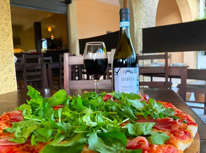 A generous herb-topped pizza alongside a bottle of red wine and a glass poured on a table in a bright dining area.