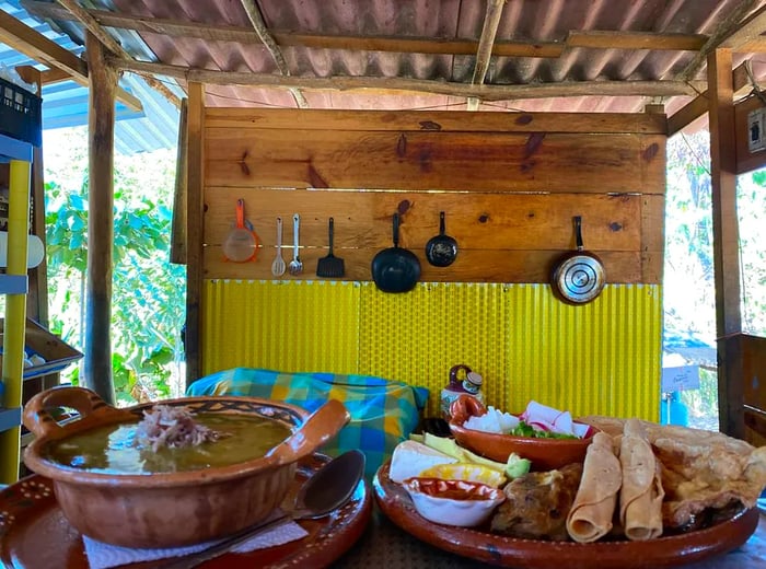 A clay bowl filled with pozole alongside a platter of tortillas and pickled vegetables, displayed in an open-air dining space adorned with pots hanging against a vibrant wall.