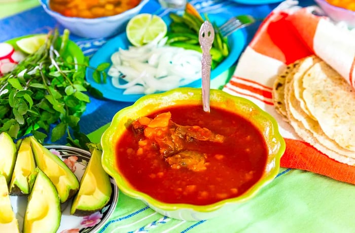 A tabletop spread featuring a bowl of mole, fresh tortillas, avocado wedges, and chopped onion and cilantro.