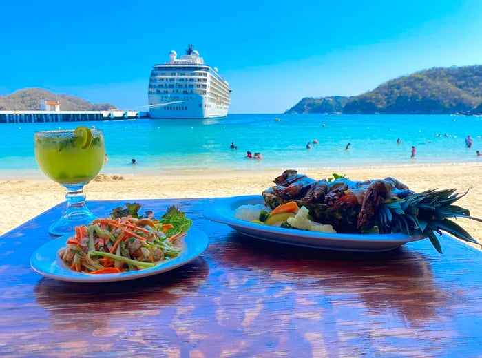 A table displays an array of dishes, including seafood-filled pineapple and a cocktail, with a beach view where a large cruise ship docks.