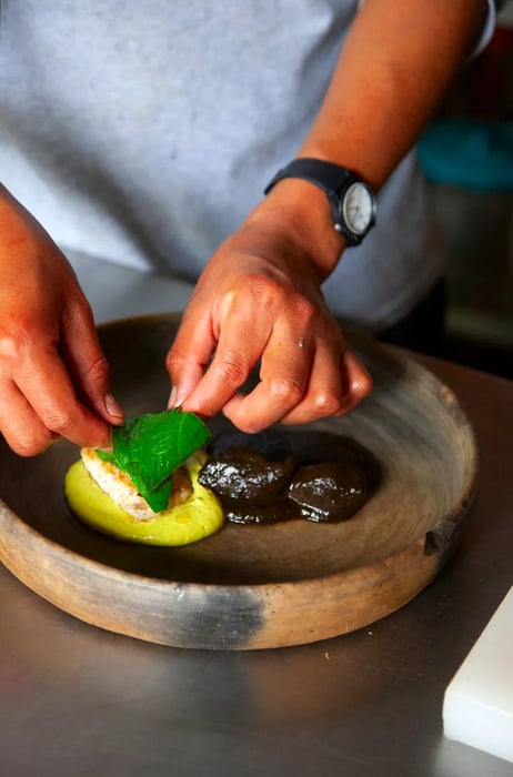 A vibrant green mole sits on a plate next to a rich brown mole, while two hands delicately place a piece of fish and a leaf atop the dish.