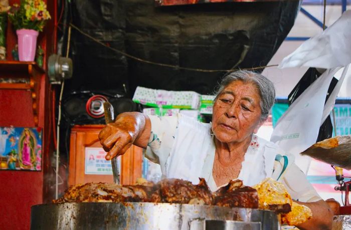 A woman stirs a large metal pot with meat resting on top.