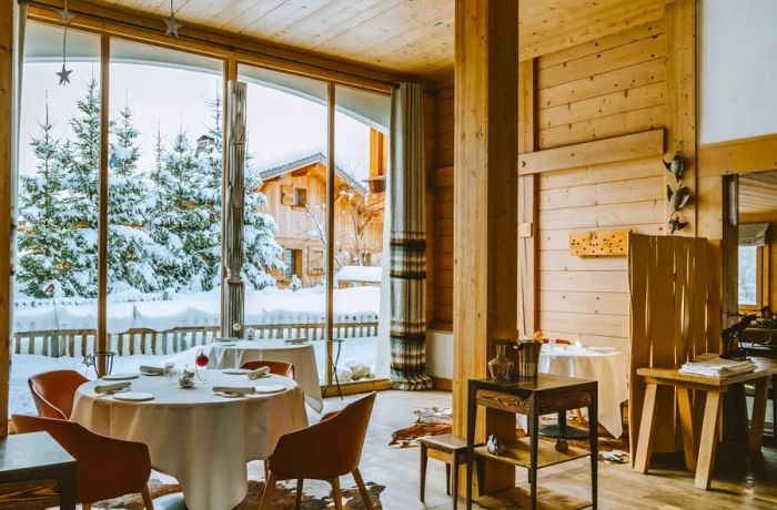 The interior of a restaurant features wooden walls and floors, large windows framing a snowy landscape, and tables adorned with white tablecloths and modern leather chairs.