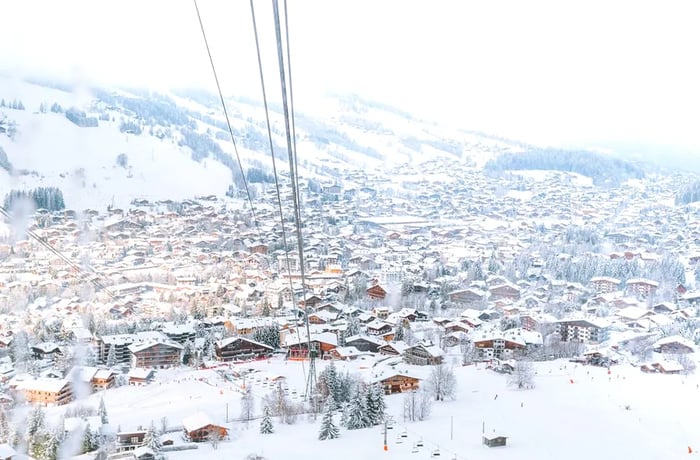 A wintry village adorned with overhead ski lift cables and towering mountains surrounding it.