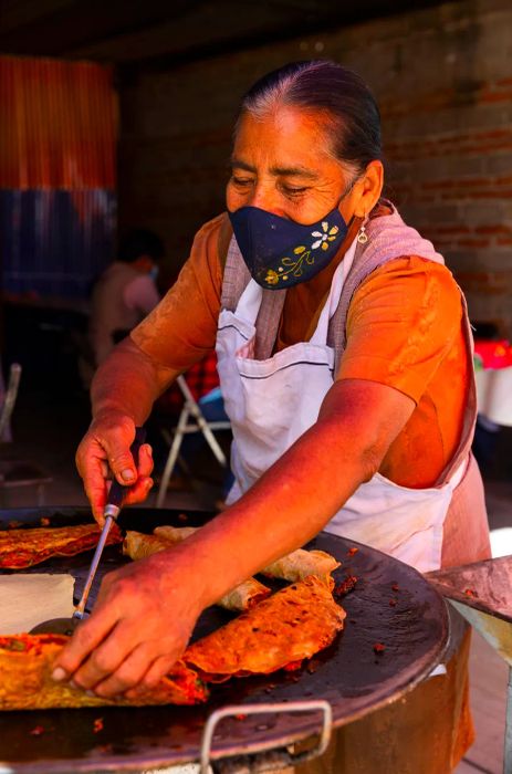 A woman in a mask skillfully flips an empanada on a comal.