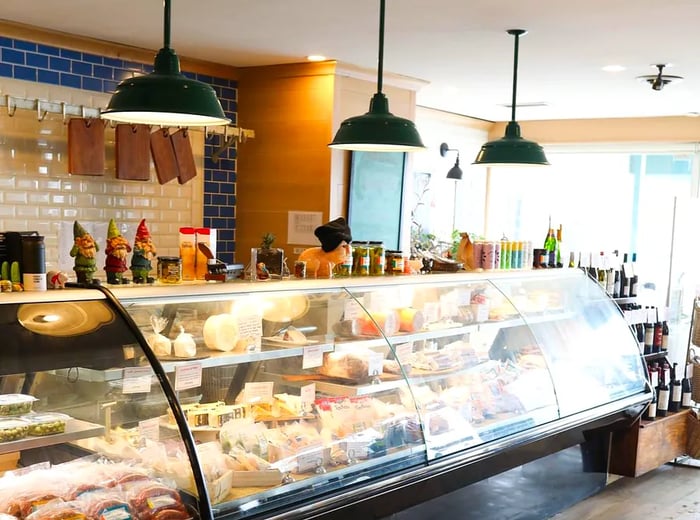 An employee stands behind a deli counter, with pendant lights illuminating the area and natural light streaming into a seating area beyond.