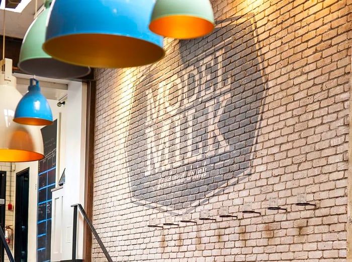 A rustic brick wall adorned with the Model Milk logo, illuminated by eggshell blue pendant lights above a long wooden table set for dining.