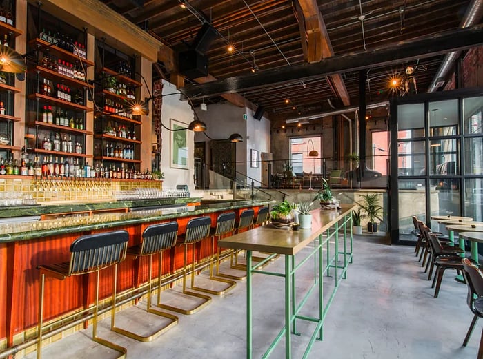 An industrial-style bar featuring soaring beamed ceilings, polished concrete floors, a wood and marble bar with leather-backed stools, a tall back bar, and cozy two-top tables by large windows.