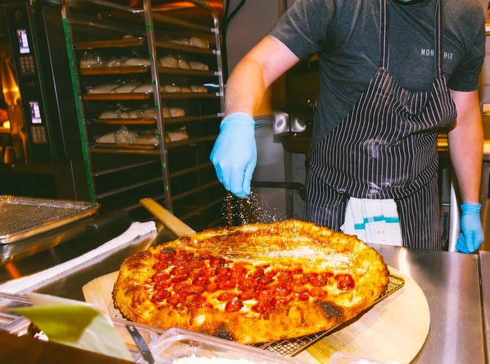 A chef wearing gloves sprinkles cheese over a pizza in the kitchen.