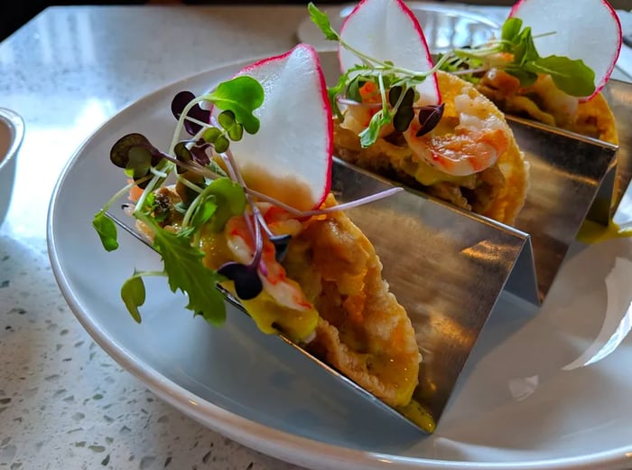 Three small tacos presented in a zigzag metal tray, topped with fresh sprouts and radish slices.