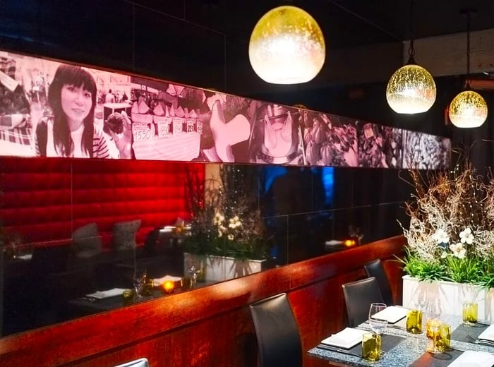 A stylish restaurant interior featuring orb pendant lights, textured tabletops, artistic plant arrangements, and a series of sepia-toned photographs adorning a black tiled wall.