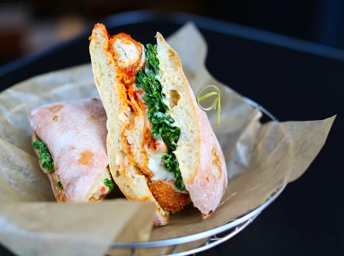 A breaded chicken sandwich loaded with greens and mayo, served on thick bread and presented in a paper-lined basket, skewered for easy handling.