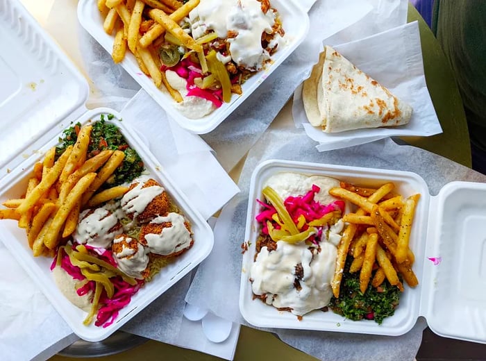 A top-down view of three plastic containers arranged on a table, filled with shawarma drizzled in sauce, pickled veggies, and crispy fries.