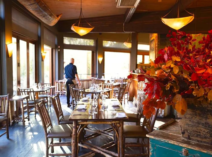 A server glides through a sun-drenched dining room, elegantly set for a meal. The space features expansive windows along one wall, vintage pendant lights, and long wooden tables.