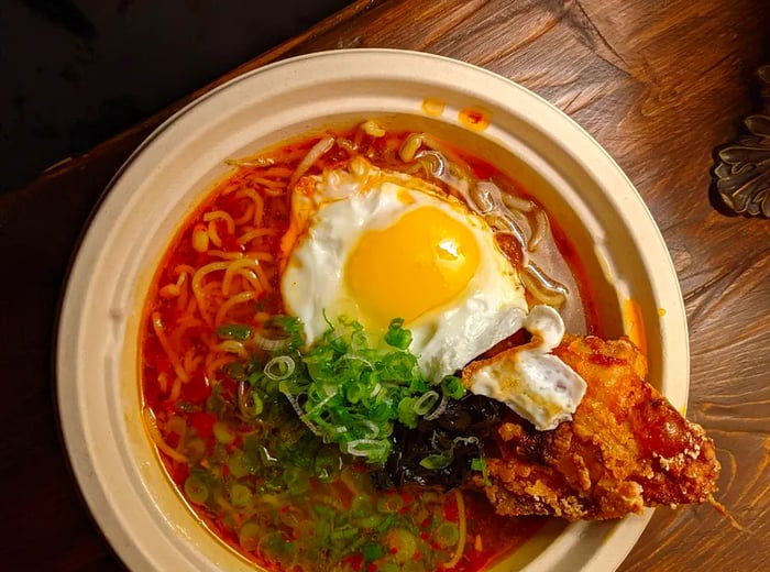 An overhead view of a light bowl filled with vibrant red soup and ramen noodles, topped with a fried egg, meat, and chopped scallions.