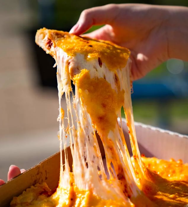 A hand lifts a cheesy slice of pizza from a box, with long strands of gooey cheese stretching behind it.