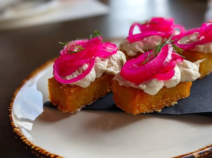 A plate featuring four large fried tater tots, generously topped with fluffy white fish and vibrant pink pickled onions.