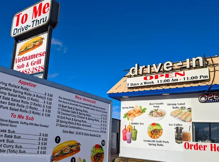 A cozy drive-thru facade featuring a menu adorned with images alongside a printed menu. A prominent sign for To Me Vietnamese Sandwiches hangs overhead in the background.