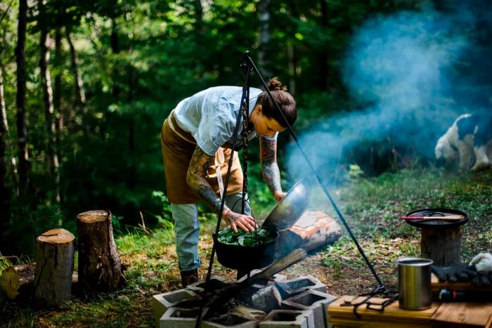 A chef prepares food over a campfire in a woodland clearing