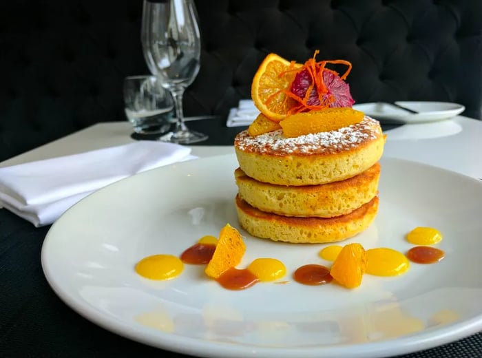 A delicate stack of airy pancakes presented on a large plate, adorned with fresh fruit, drizzles of sauce, and topped with additional candied fruit slices.