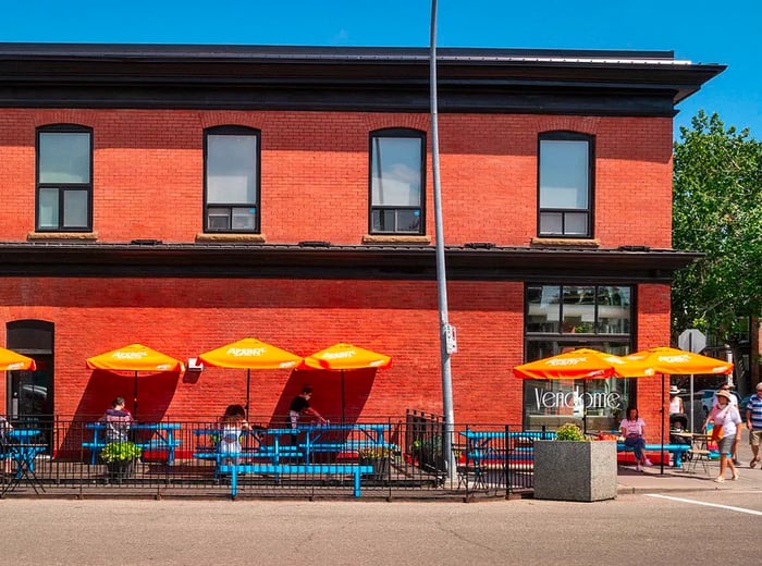 A long red brick building exterior features a sidewalk patio adorned with bright yellow umbrellas, where customers can be seen sitting and strolling nearby.