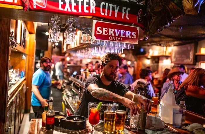 A bartender looks at the camera while reaching for glassware, with a lively bar scene in the background.