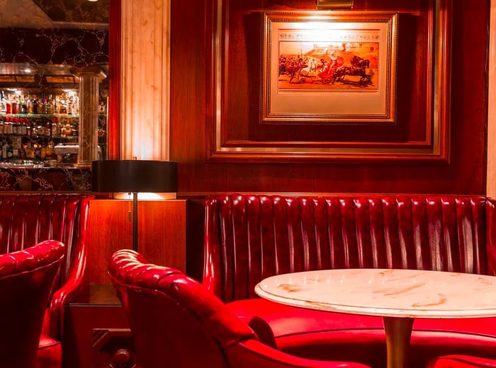 A plush red leather booth accompanied by thick leather armchairs around a marble table. On the dark wooden wall hangs an illustration of a horse race, while a dark marble bar awaits around the corner.