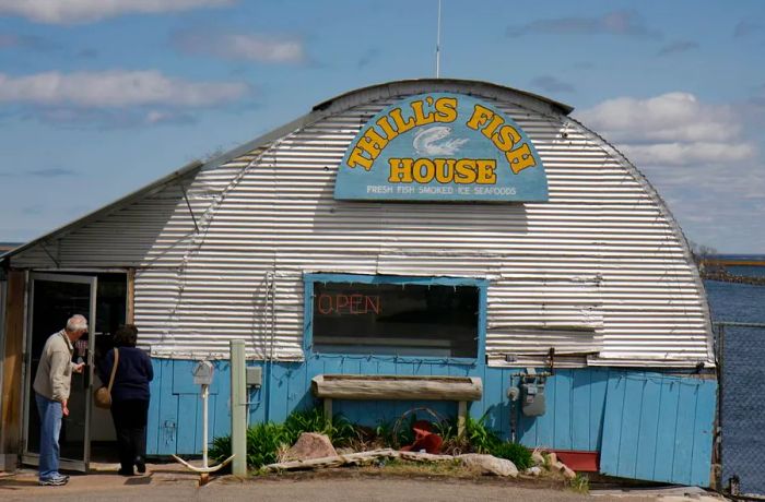 A customer holds the door for another person entering a rounded metal building adorned with slatted siding and a sign featuring a leaping fish, reading ‘Thill’s Fish House fresh fish smoked ice seafoods’