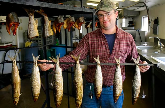 A man holds a long line of skewered fish in front of fish pieces drying in a kitchen.