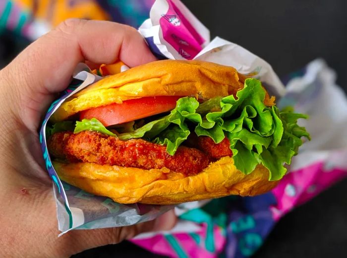 A hand grasps a compressed sandwich filled with imitation fried chicken, lettuce, and tomato, wrapped in foil.