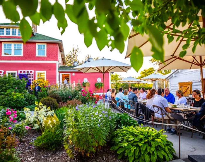 A group of diners enjoys a long communal table nestled among trees and shrubs, in front of a spacious farmhouse-style establishment.