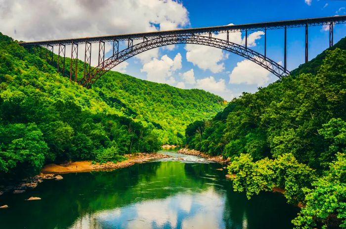 The New River Gorge Bridge viewed from Fayette Station Road on a partly cloudy day, framed by lush greenery on both banks