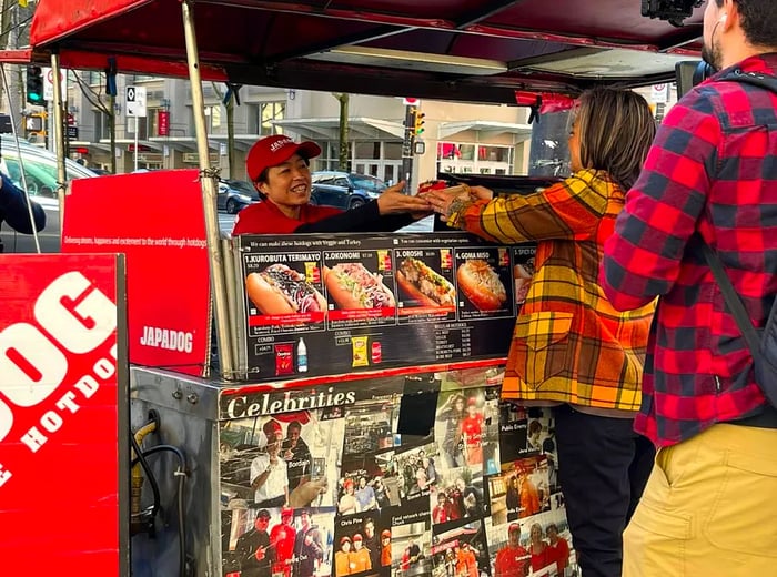 A vendor serves a hot dog to customers from a food cart displaying a variety of menu options.