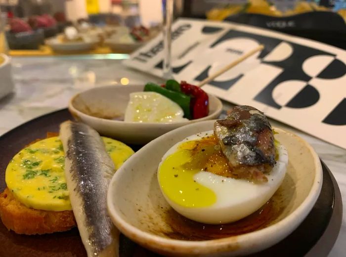 A close-up of a boiled egg topped with sardine, a small fried patty adorned with herb butter and another fish, and a skewer with additional treats, all resting on a marble countertop.