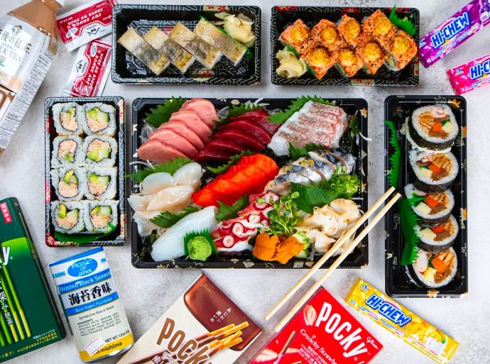 Takeout boxes filled with maki rolls, sashimi, and kimbap, accompanied by snacks like Pocky and Hi-Chew, arranged on a textured surface.