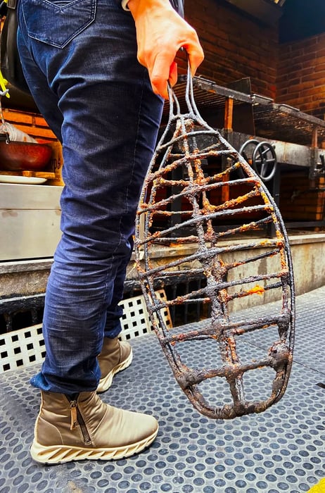 A person grips a long, fish-shaped metal basket in front of a grill