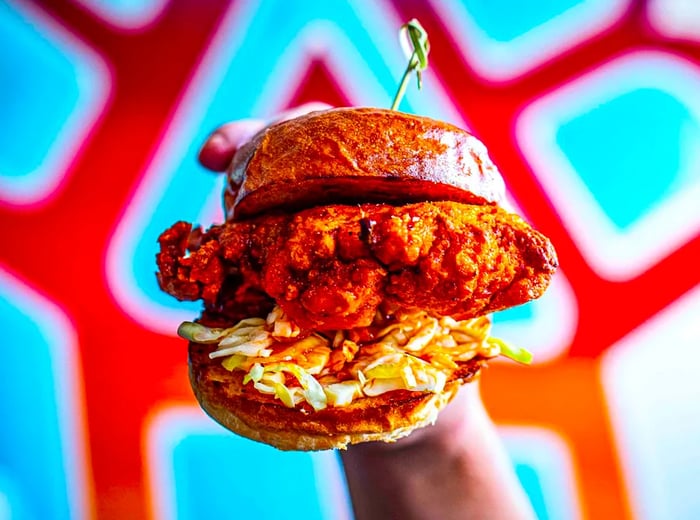 A hand presenting a generously filled fried chicken sandwich against a vibrant backdrop