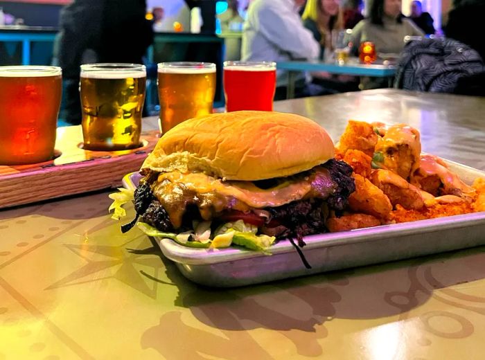 A wide, flat burger adorned with sauce and lettuce, served on a quarter sheet alongside fries and a flight of beers.