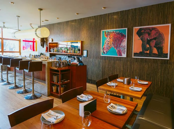 The restaurant features a cozy interior with a textured wood wall, a bar illuminated by a long pendant light, and tables elegantly set with place settings, adorned with illustrations of a zebra and elephant.