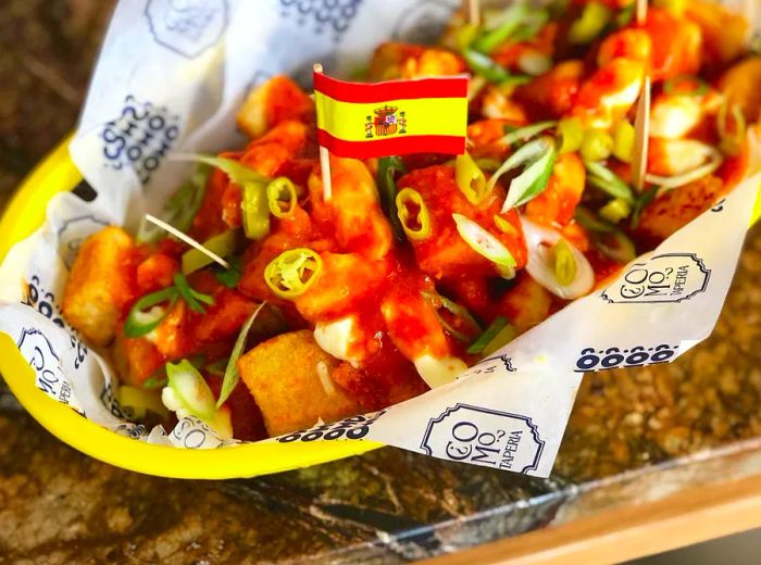 A basket lined with wax paper overflowing with poutine, adorned with slices of peppers and sauces, and topped with a small Spanish flag.