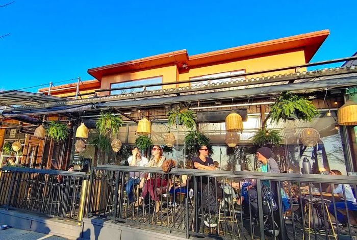 Diners enjoy their meals on a patio outside a quaint building, shaded by a trellis adorned with hanging lanterns.