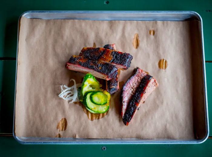 A metal platter lined with butcher paper, featuring a selection of barbecued ribs, brisket slices, and pickles