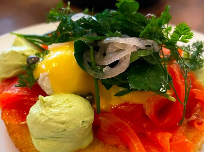 A close-up of a rosti topped with slices of smoked salmon, pickled vegetables, greens, and a boiled egg.