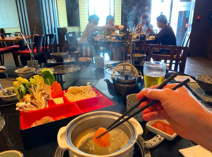 A diner holds a piece of fish on chopsticks, dipping it into boiling water while seated alone at a table.