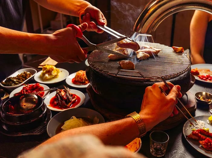 Hands wield tongs and scissors to cut meat over a grill, while other pieces roast nearby, as diners enjoy a table filled with banchan.