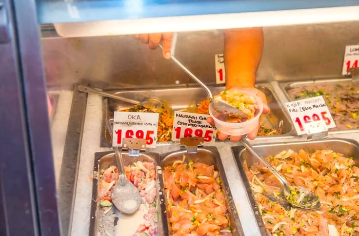 Through the glass of a deli case, a worker scoops poke into a plastic container.