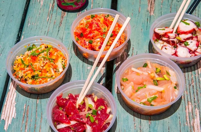 Five varieties of poke in plastic containers are arranged on a blue picnic table, with chopsticks poking out of several.