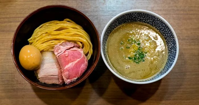 An aerial view of two bowls: one filled with thick noodles, slices of pork, and a dark boiled egg, while the other showcases a rich, thick green sauce.