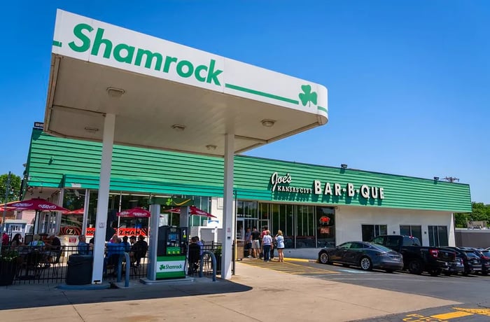 The exterior of a restaurant located within a Shamrock gas station, with the name Joe’s Kansas City Bar-B-Que. Diners are queuing outside.